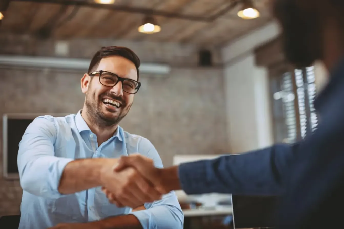 Smiling businessman in a button-down shirt shaking hands with a partner during a successful professional meeting in a modern office.