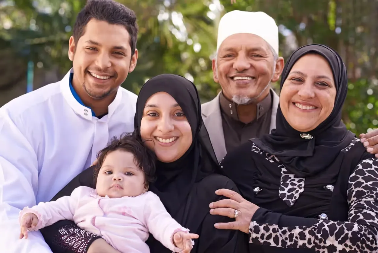 A smiling family of five, including two women in black hijabs, a man in a white cap, a man in a white shirt, and a baby in a pink outfit, poses together outdoors with greenery in the background.
