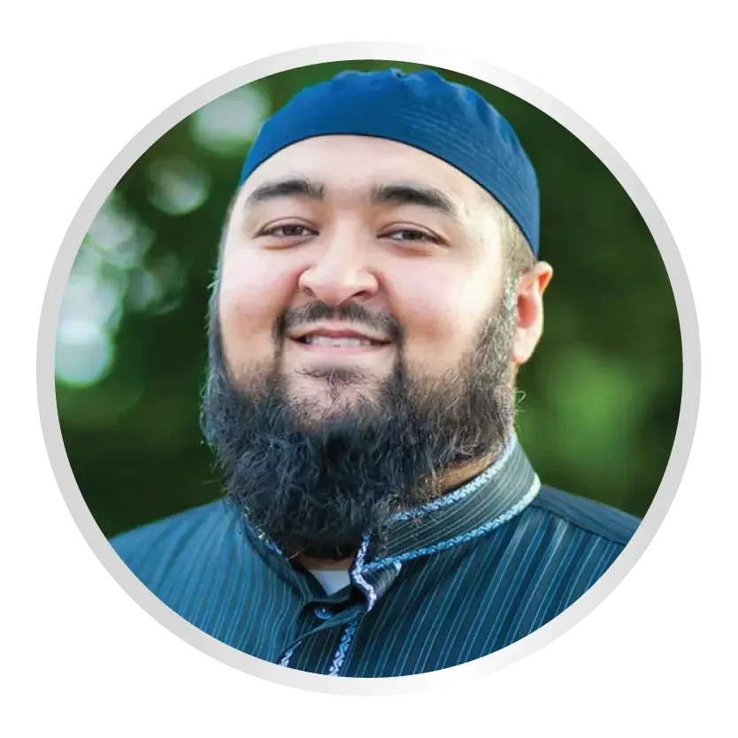 A smiling man with a full beard wears a dark blue kufi cap and a striped, collared shirt against a soft, green outdoor background.
