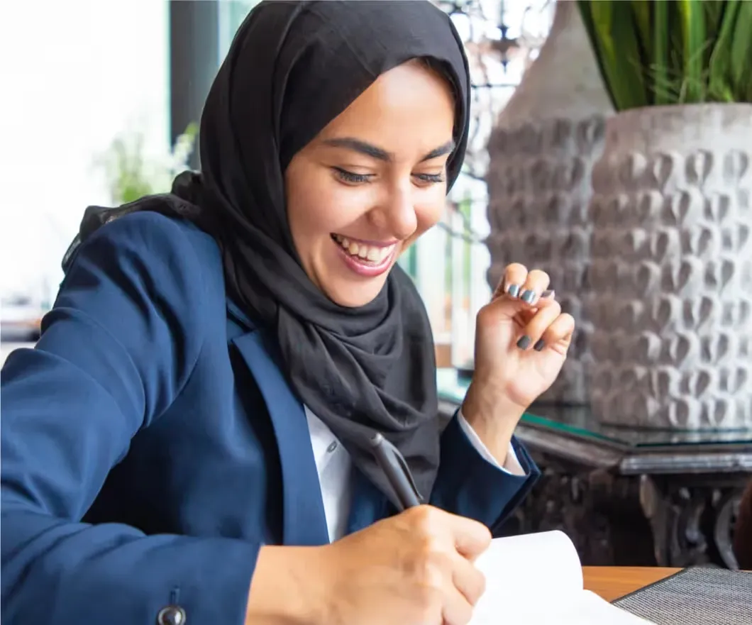 A smiling woman wearing a black hijab and a blue blazer writes in a notebook while sitting at a table.