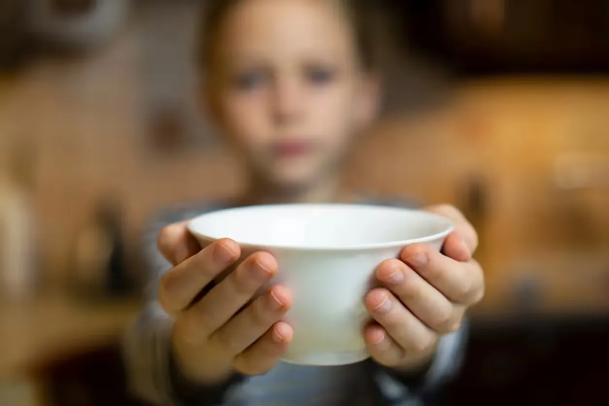 A child holds out an empty white ceramic bowl with both hands, while their face and the warm-toned background remain softly out of focus.