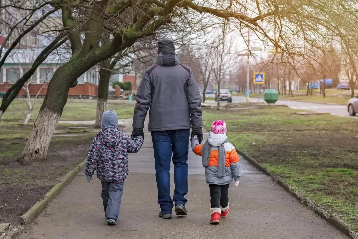 A man and two young children, seen from behind, hold hands as they walk down a paved path on a cool day. They are dressed in warm winter coats and hats, surrounded by bare trees and a quiet street in the background.