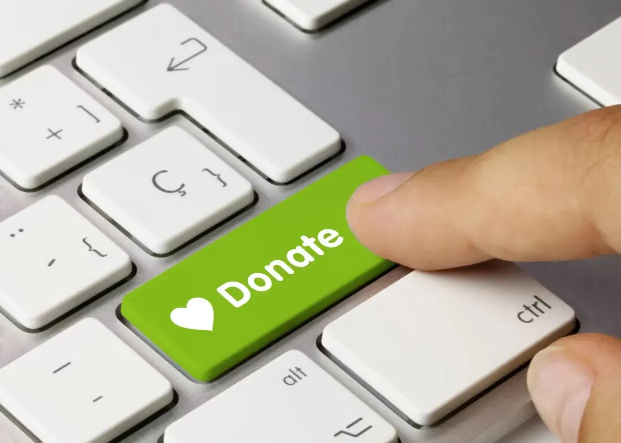 A finger presses a bright green key on a computer keyboard, which features a white heart icon and the word "Donate."