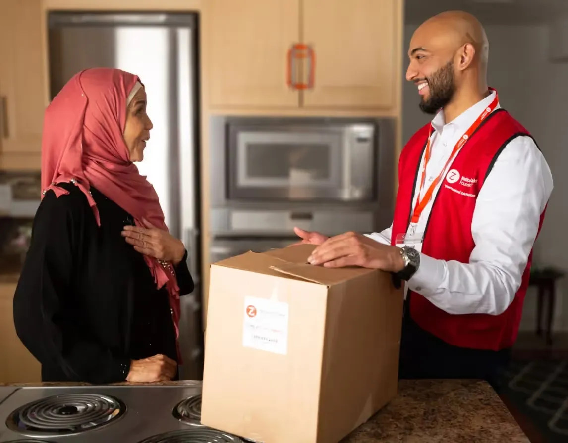 A smiling man wearing a red National Zakat Foundation vest delivers a cardboard box to a woman wearing a pink hijab in her kitchen. The woman looks at him warmly with her hand placed over her chest.