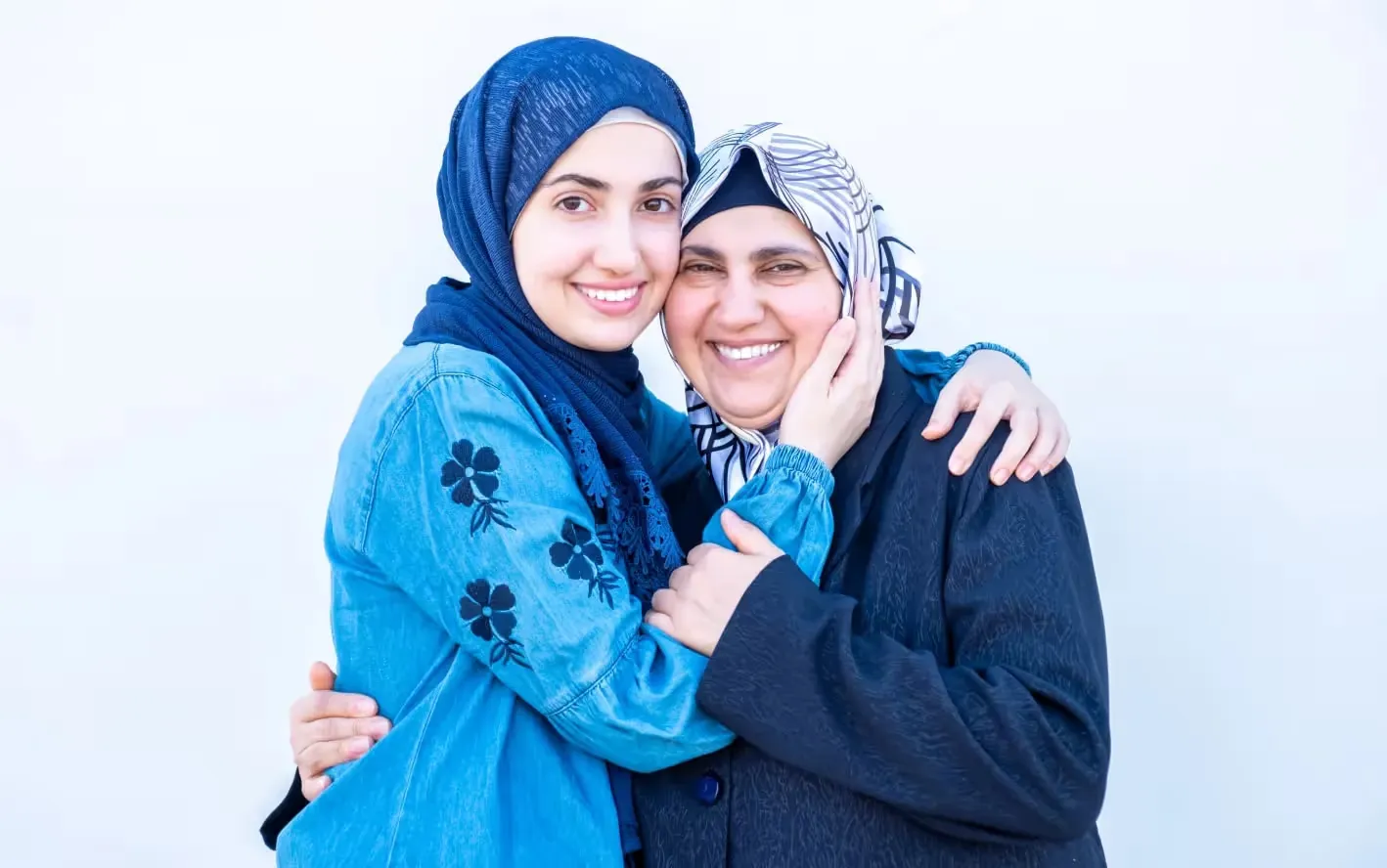 Two smiling women wearing hijabs embrace closely against a plain, light background. The younger woman on the left wears a blue hijab and denim shirt, gently cupping the cheek of the older woman on the right, who wears a patterned hijab and dark jacket.