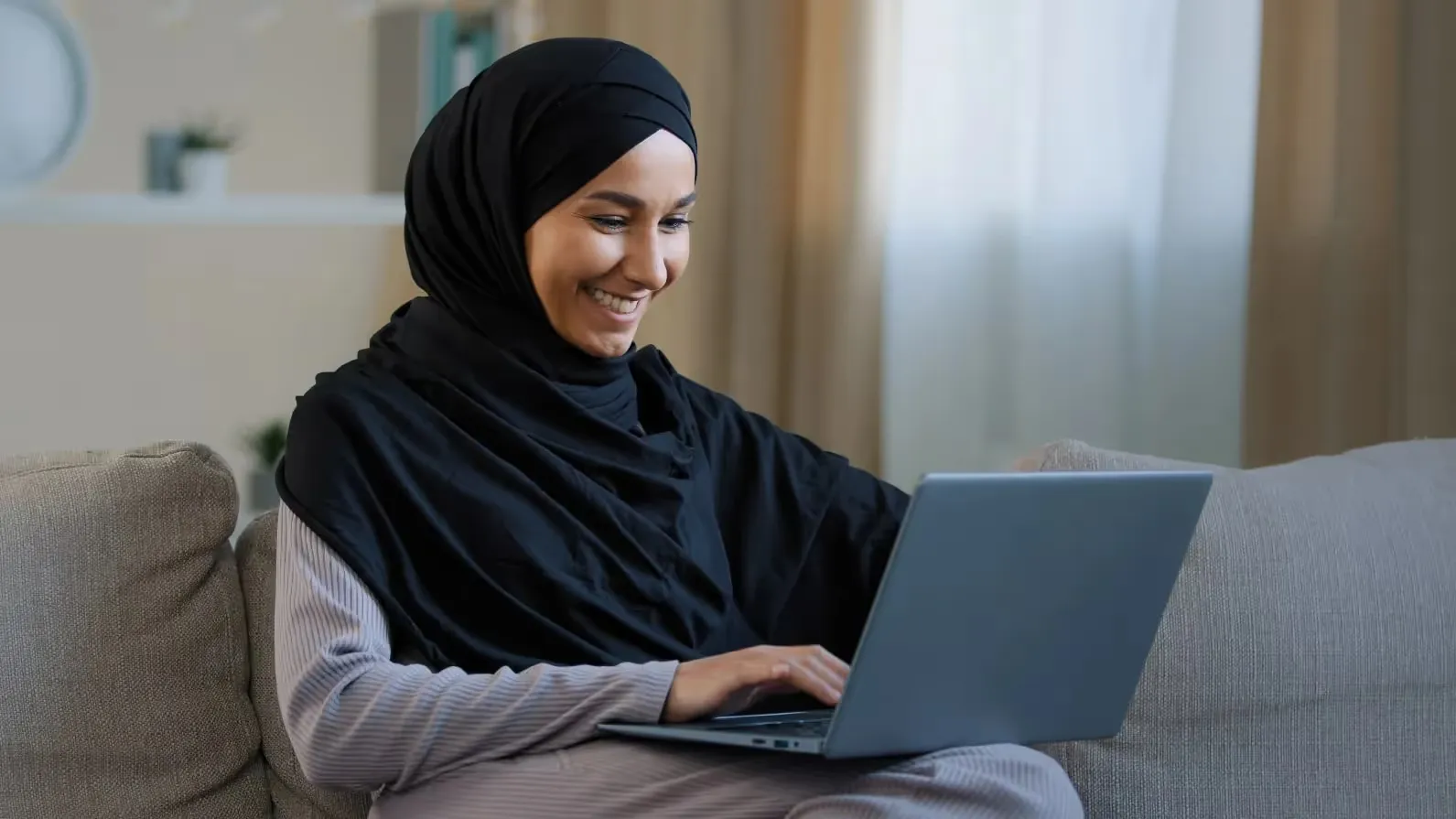 A smiling woman wearing a black hijab sits on a grey couch while typing on a laptop. The background shows a softly blurred living room with natural light filtering through a window.