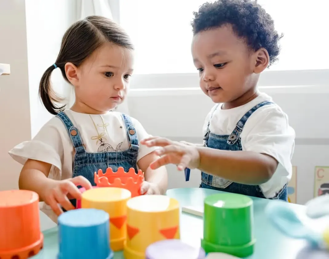Two toddlers, a girl with a ponytail and a boy with curly hair, wear matching denim overalls while playing with colorful plastic stacking cups at a table.