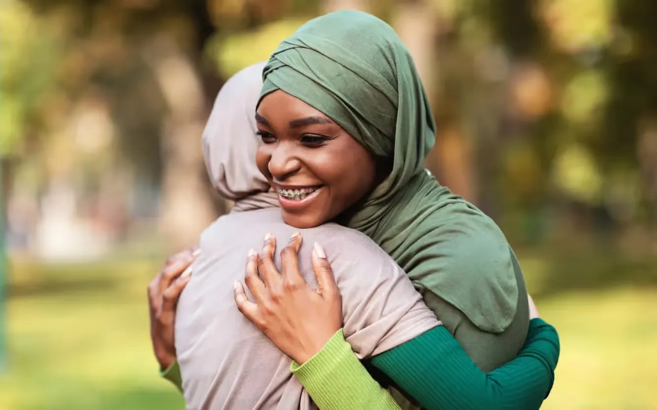 A smiling Black woman wearing a green hijab and dental braces warmly embraces another woman wearing a beige hijab in an outdoor park with a blurred green background.