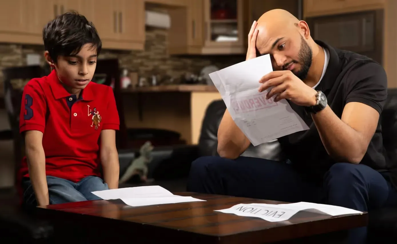 A man sits at a table with a young boy, looking distressed while holding a document that clearly displays the word "EVICTION" in large red letters. Another paper with the same "EVICTION" text lies on the table in front of them.