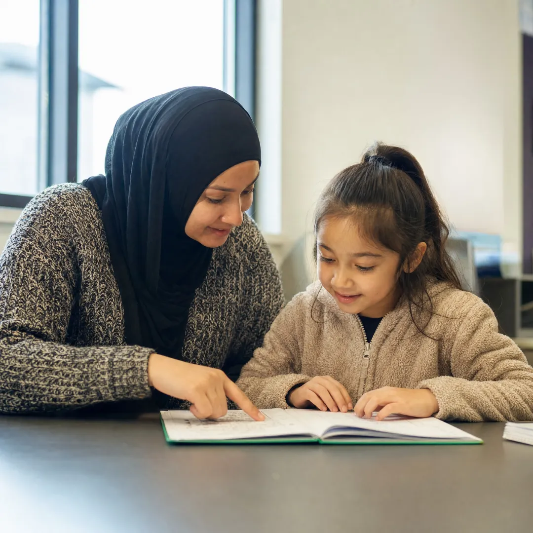 Woman in a black hijab points to a page as a young girl reads beside her, sharing a warm, focused learning moment over an open book.