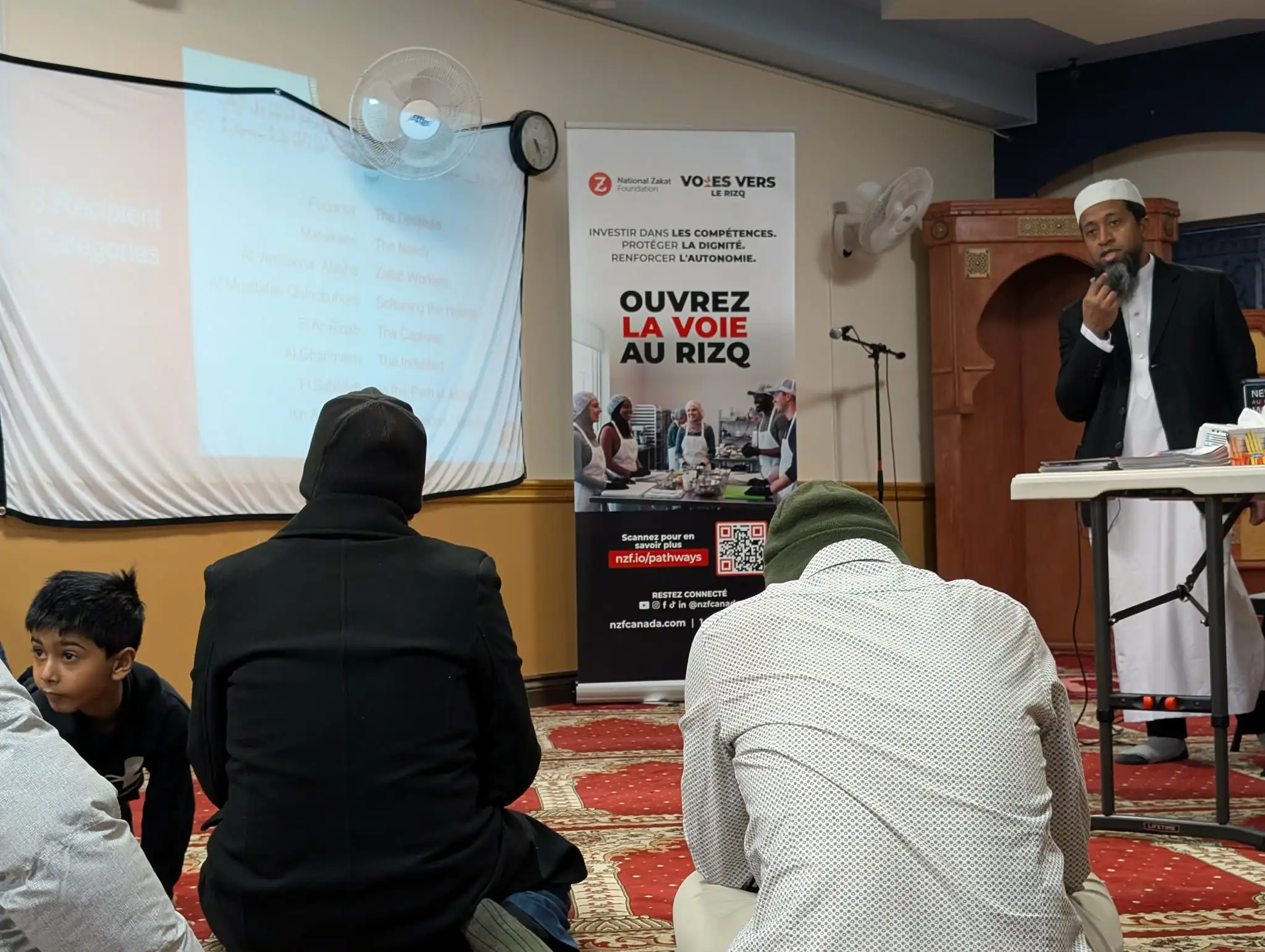 A Muslim speaker addresses a seated audience in a mosque, beside a projected presentation and French-language banner; the gathering feels calm and attentive.