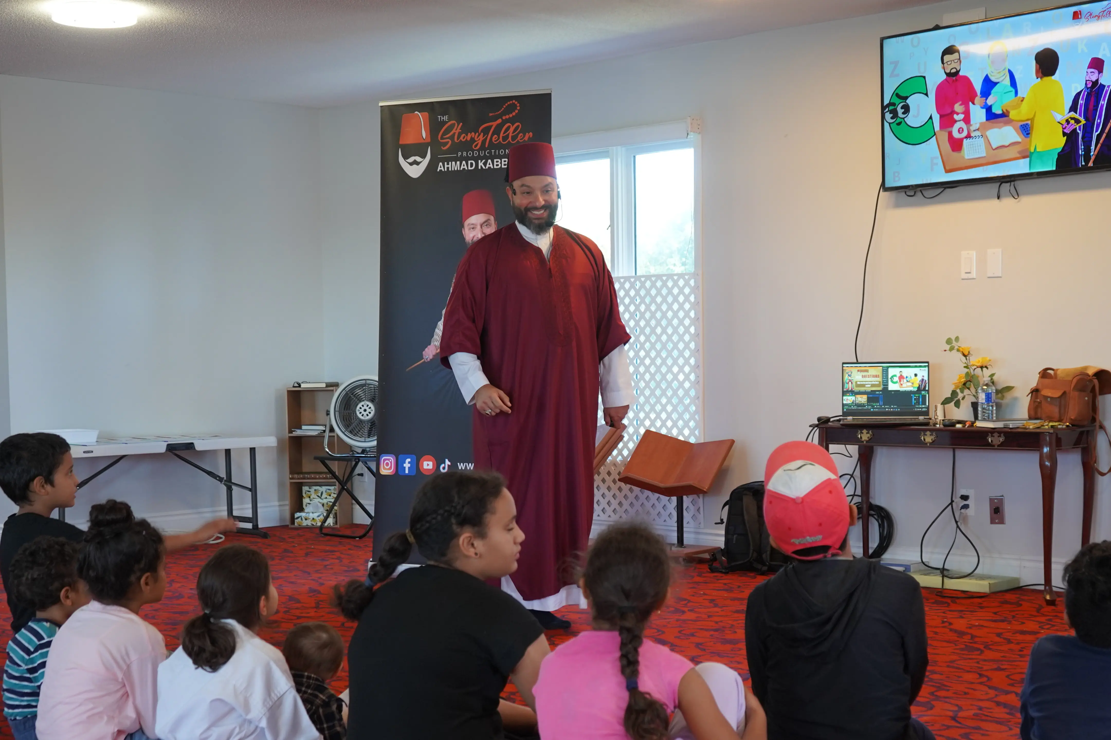A smiling man in a maroon robe warmly addresses children seated on a red carpet in a bright room, while a cartoon plays on a wall-mounted TV.