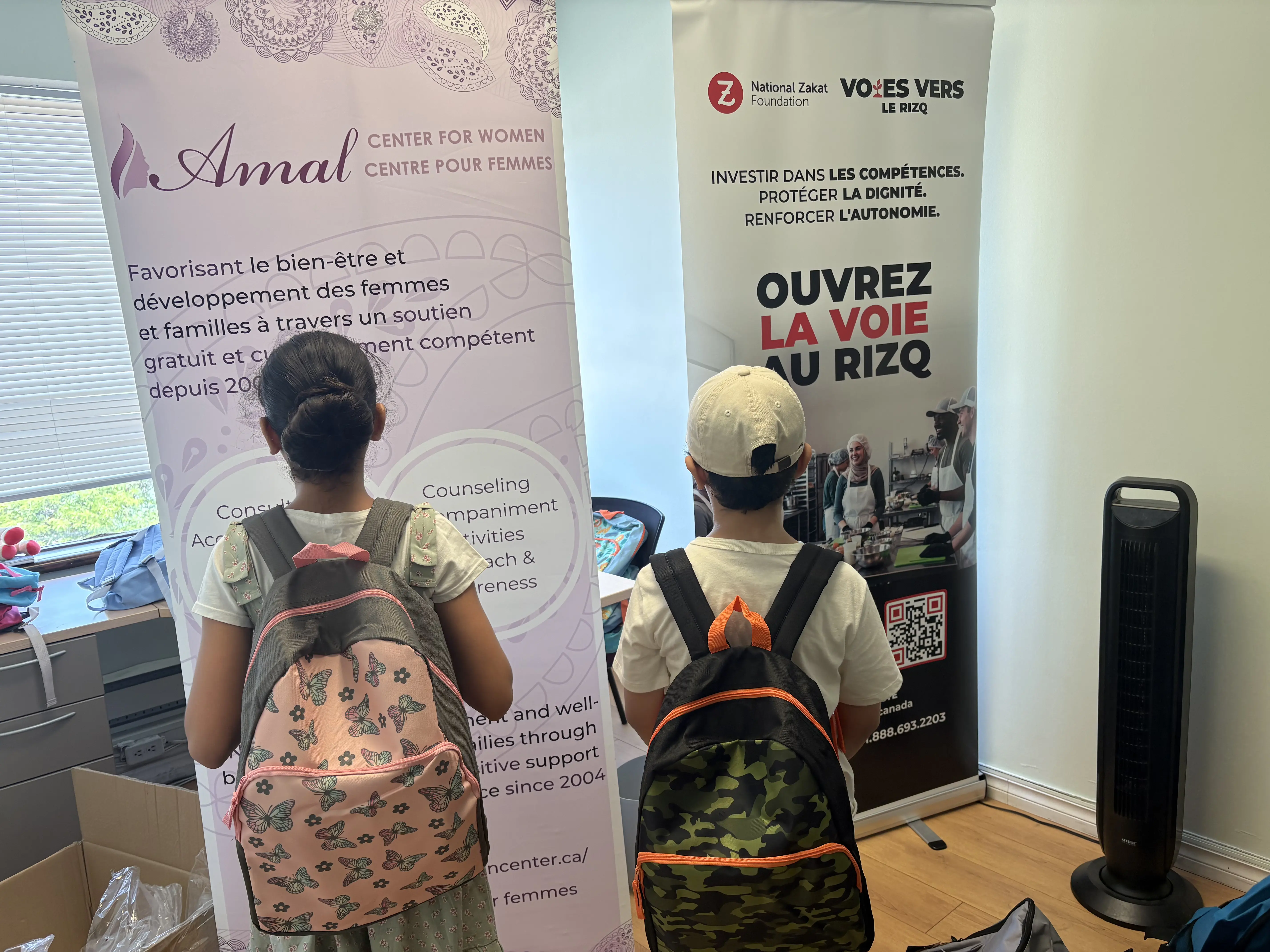 Two visitors with pink floral and camouflage backpacks face French banners promoting women’s support and youth empowerment at an indoor event.