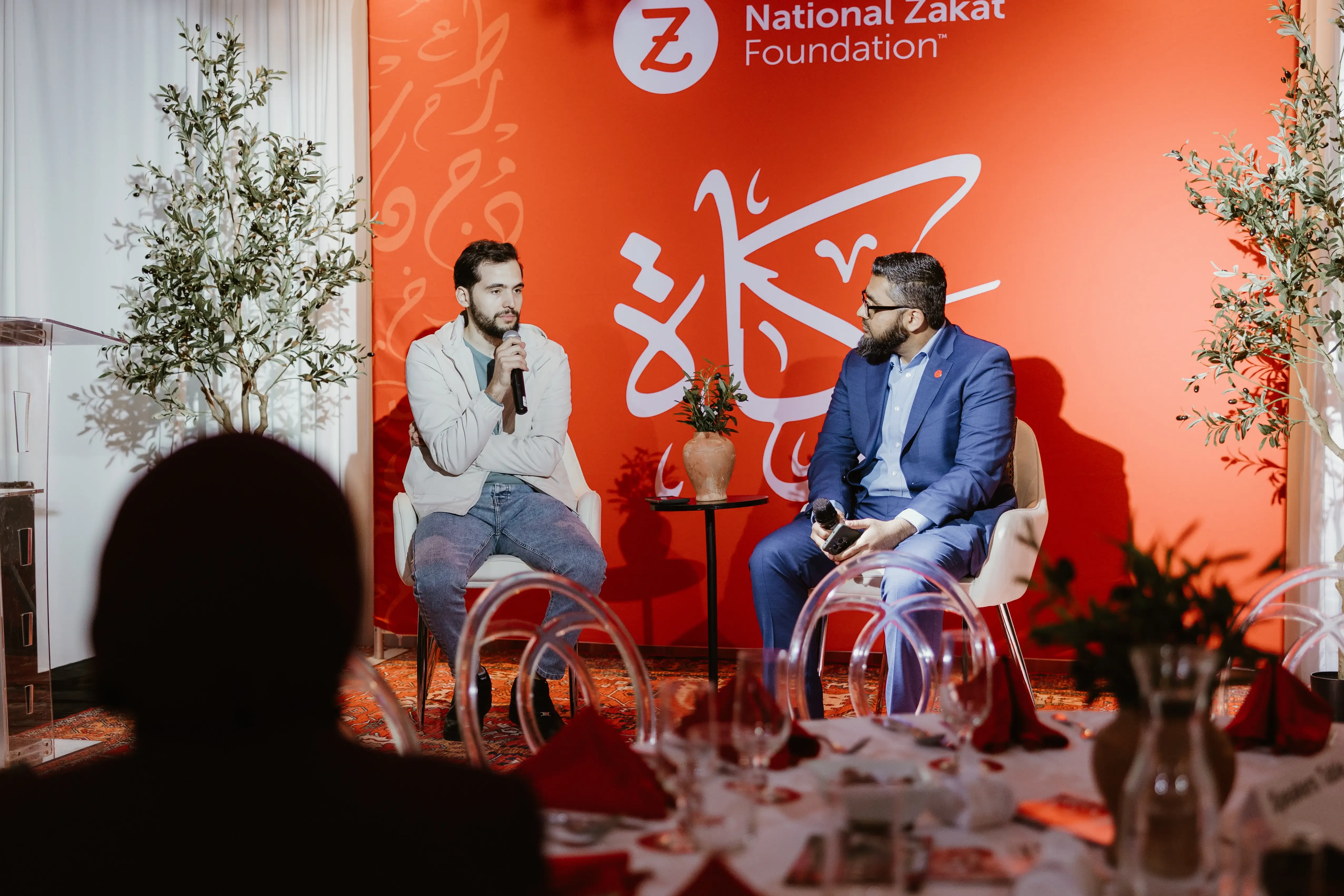 Two men speak onstage before a red National Zakat Foundation backdrop, seated beside a small table; audience silhouettes and banquet tables fill the foreground.