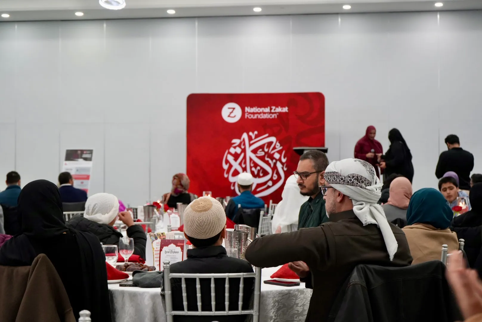 Guests share a meal beneath a National Zakat Foundation banner; diverse attendees in modest dress create a warm, charitable gathering.