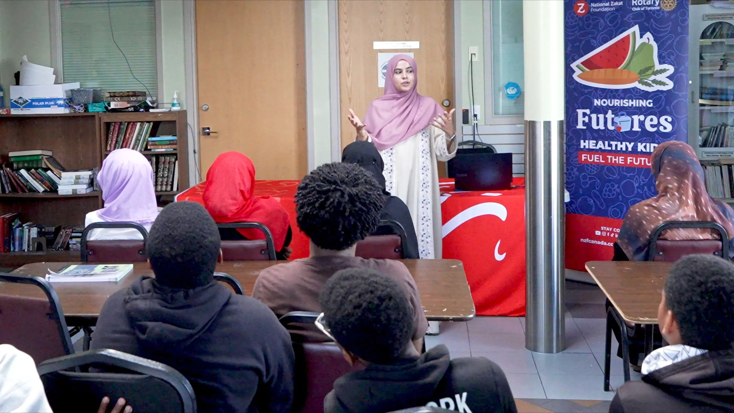 A woman in a pink hijab speaks to seated students in a classroom, beside a “Nourishing Futures” nutrition poster; the scene feels educational and community-focused.