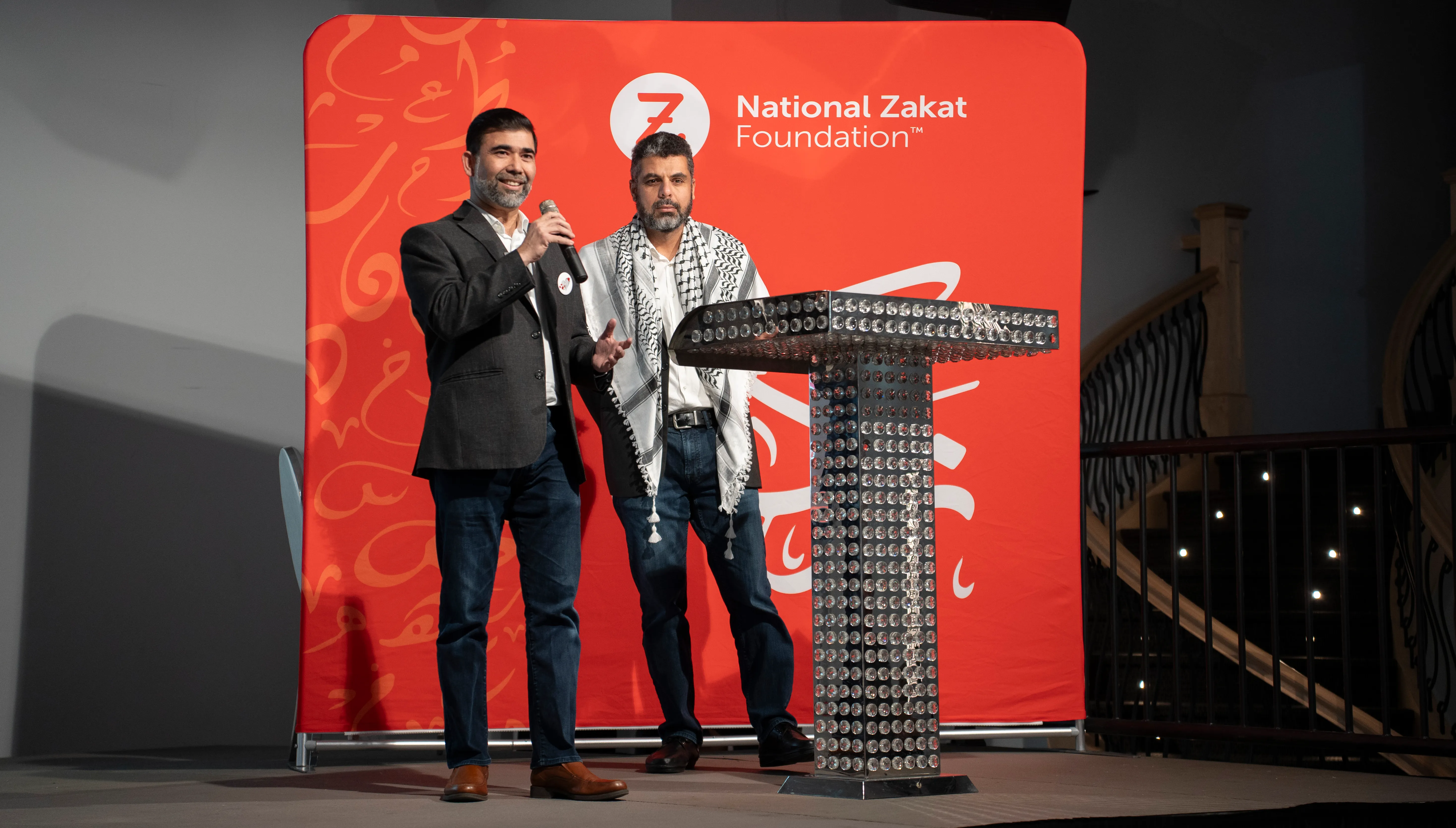 Two men stand beside a jeweled podium onstage before a red National Zakat Foundation backdrop; one holds a microphone, creating a formal event atmosphere.