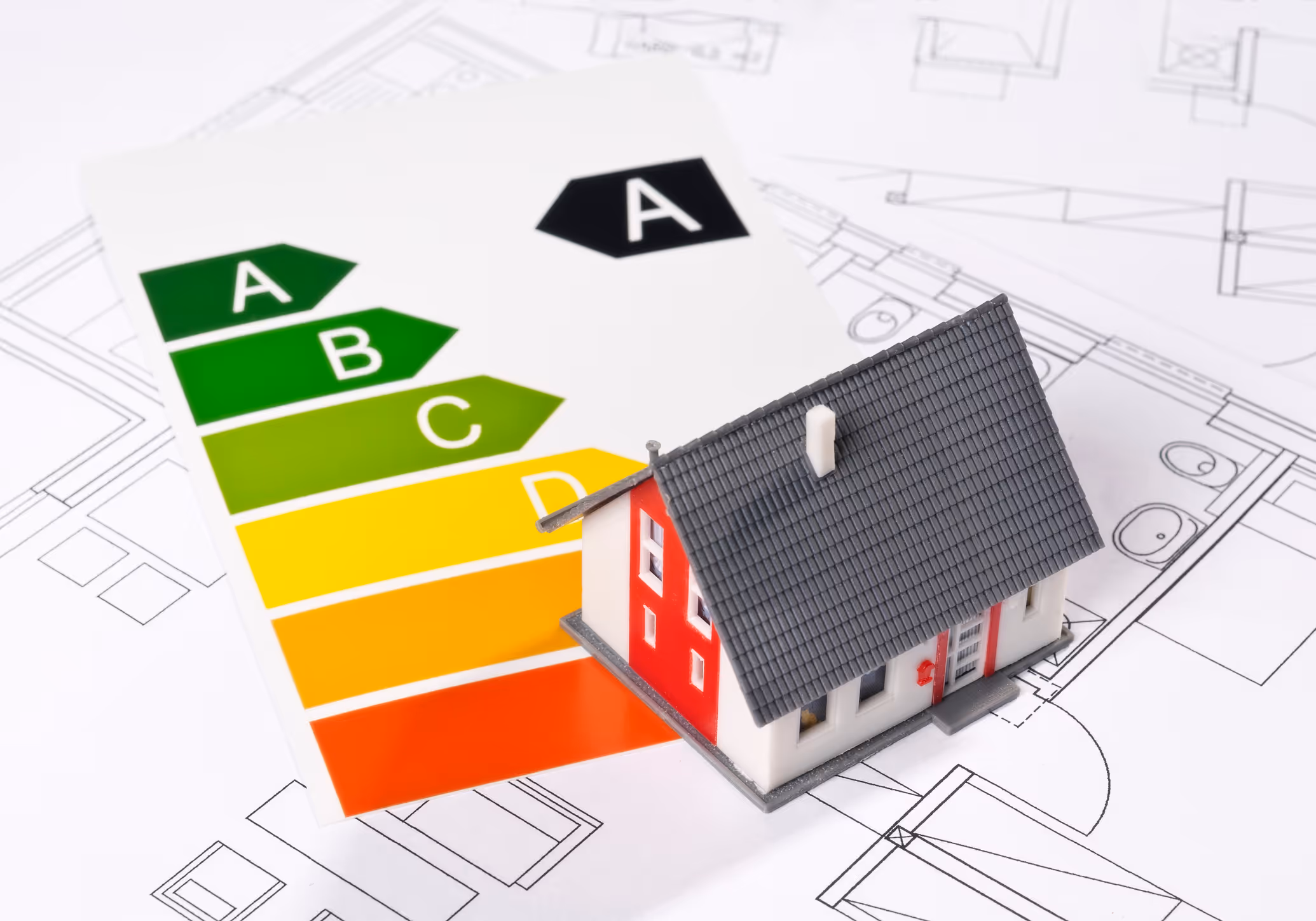 Rot-weißes Miniaturhaus steht auf dem Tisch auf Grundrisszeichnungen und einer energetischen Bewertungsskala mit den Effizienzklassen A (grün) bis F (rot).