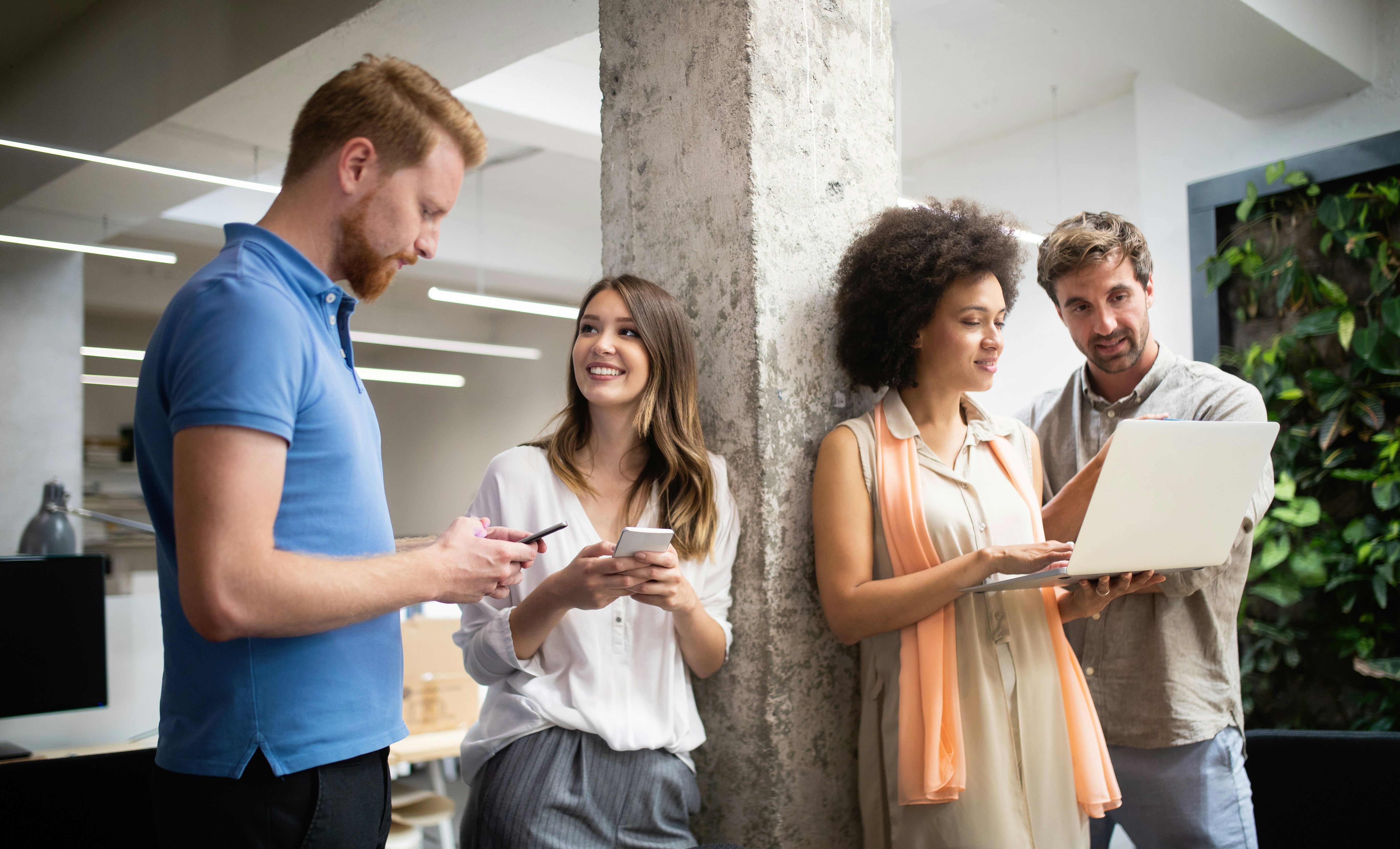 Vier Kollegen stehen mit Smartphones und Computer in einer Wohnung die zum modernen Büro umgewandelt wurde.