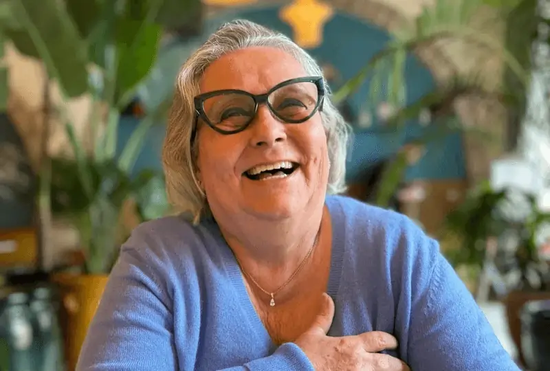 Photo de Chantal, 73 ans, souriante et sereine, la main sur le cœur, assise à une table colorée.