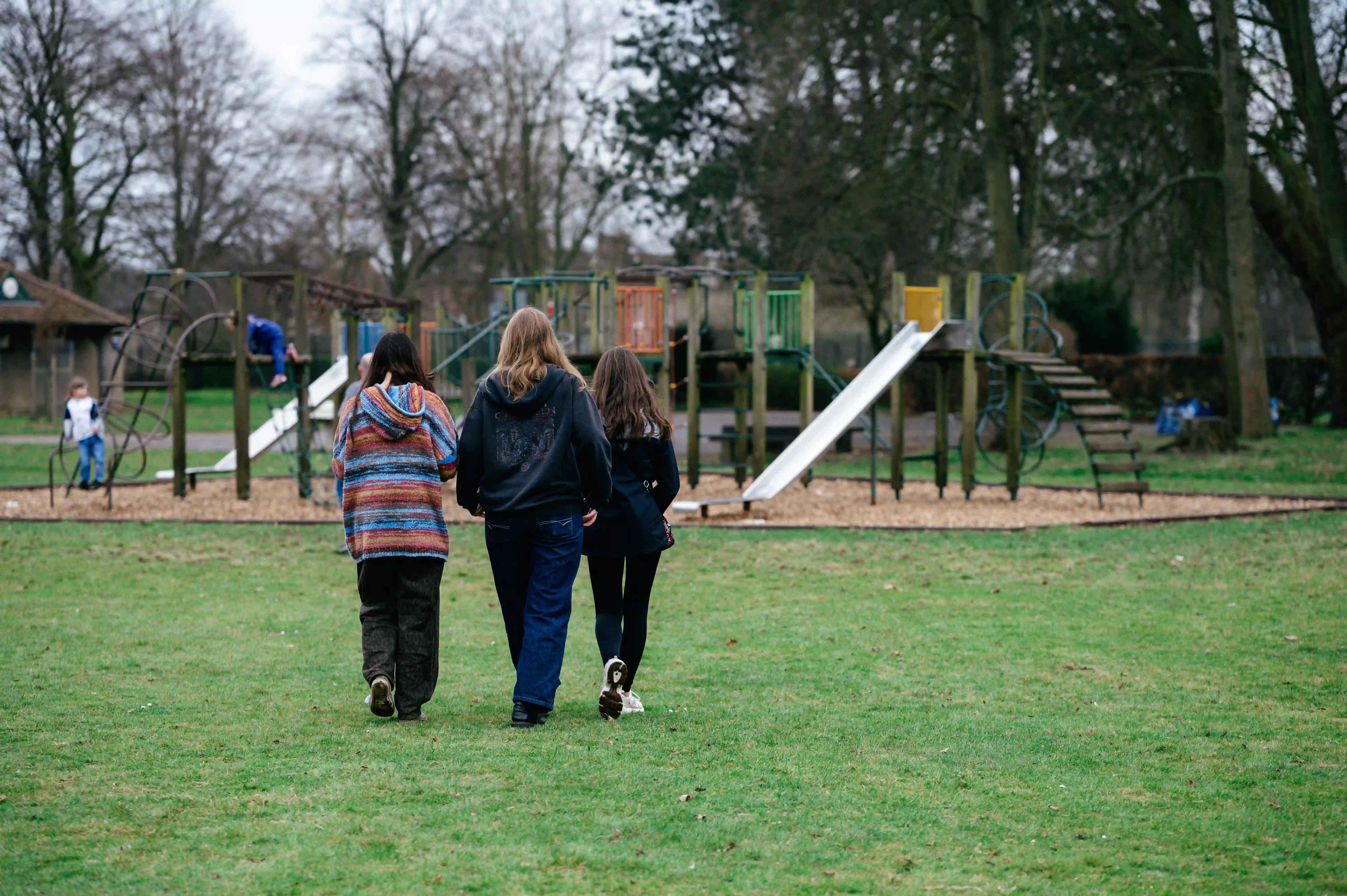 Three girls walking toward a playground with slides and climbing structures on a grassy field.
