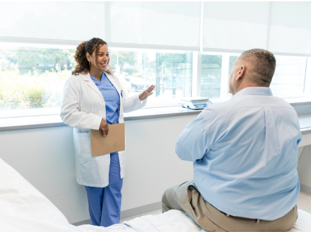 Clinician in a white coat and blue scrubs talks with a seated patient in a bright exam room, holding a clipboard while gesturing
