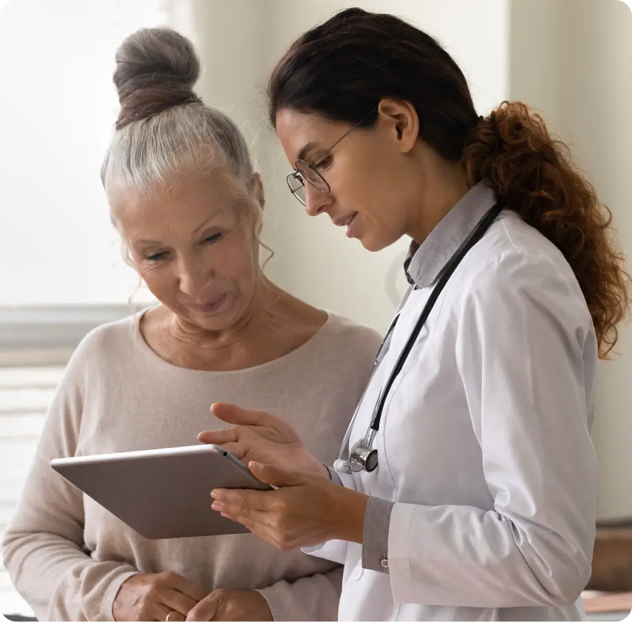 Female doctor showing a tablet screen to an elderly woman in a light room.
