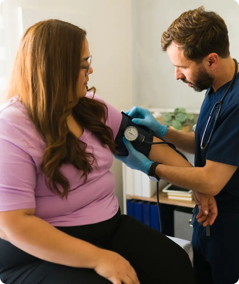 Healthcare professional measuring blood pressure of seated woman wearing a purple shirt.