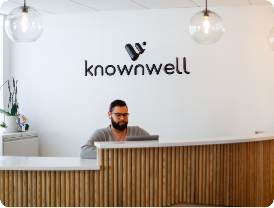 Man working on laptop behind a wood-paneled reception desk with Knownwell logo on white wall.