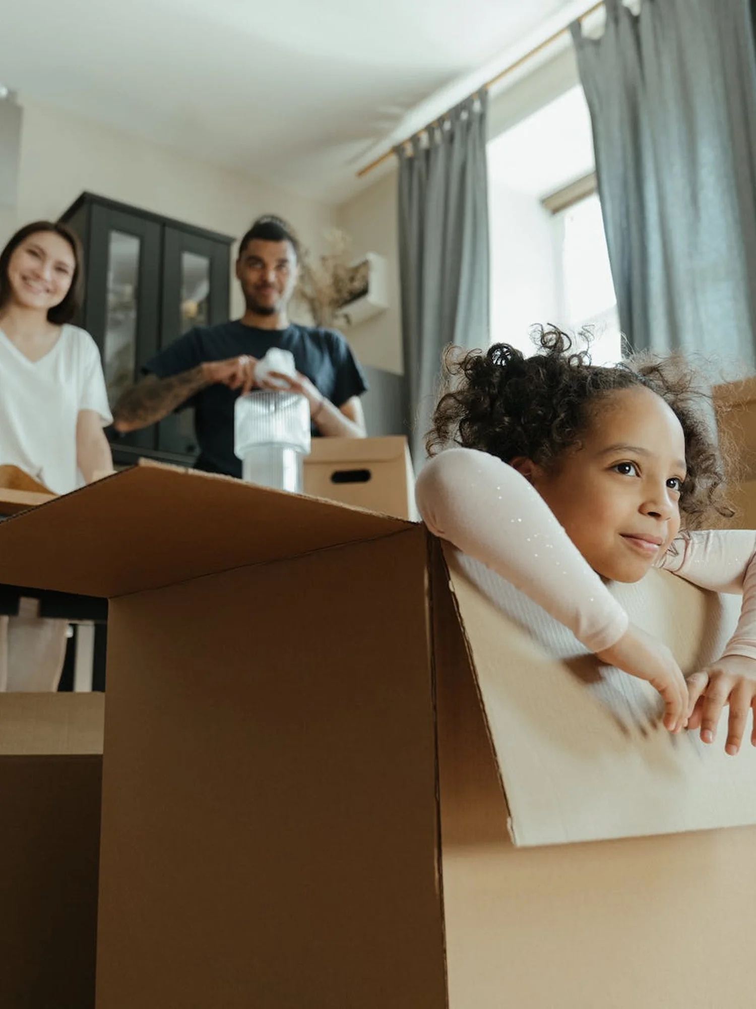 A little girl is peeking out from the a box with a smile and a man is standing behind out of focus