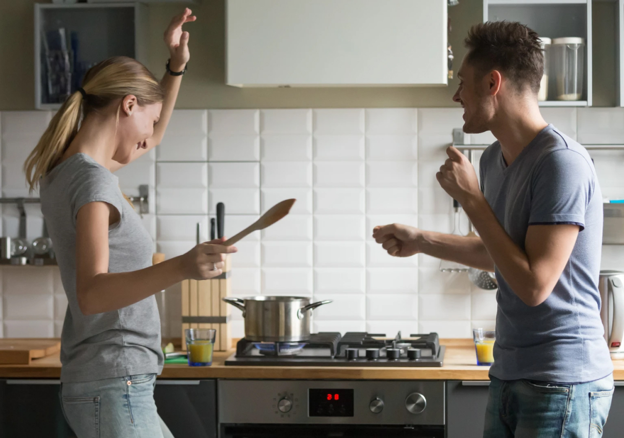 A young couple enjoys cooking together in a modern kitchen, smiling and dancing playfully near the stove with a pot on the burner and wooden utensils in hand.