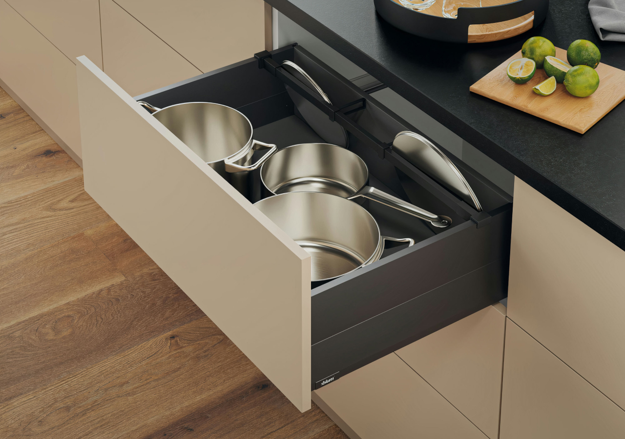 Open deep kitchen drawer with organized stainless steel pots and lids, matte beige fronts, and black countertop.