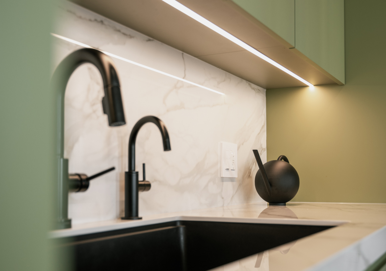 Close-up of a modern kitchen sink with matte black faucets, marble-look backsplash, and green cabinets with under-shelf lighting.
