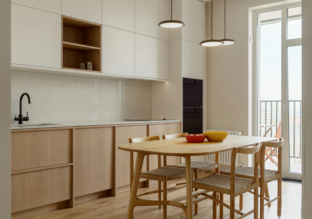 Bright minimalist kitchen with light oak cabinets, white uppers, black fixtures, and a wooden dining set by large windows.