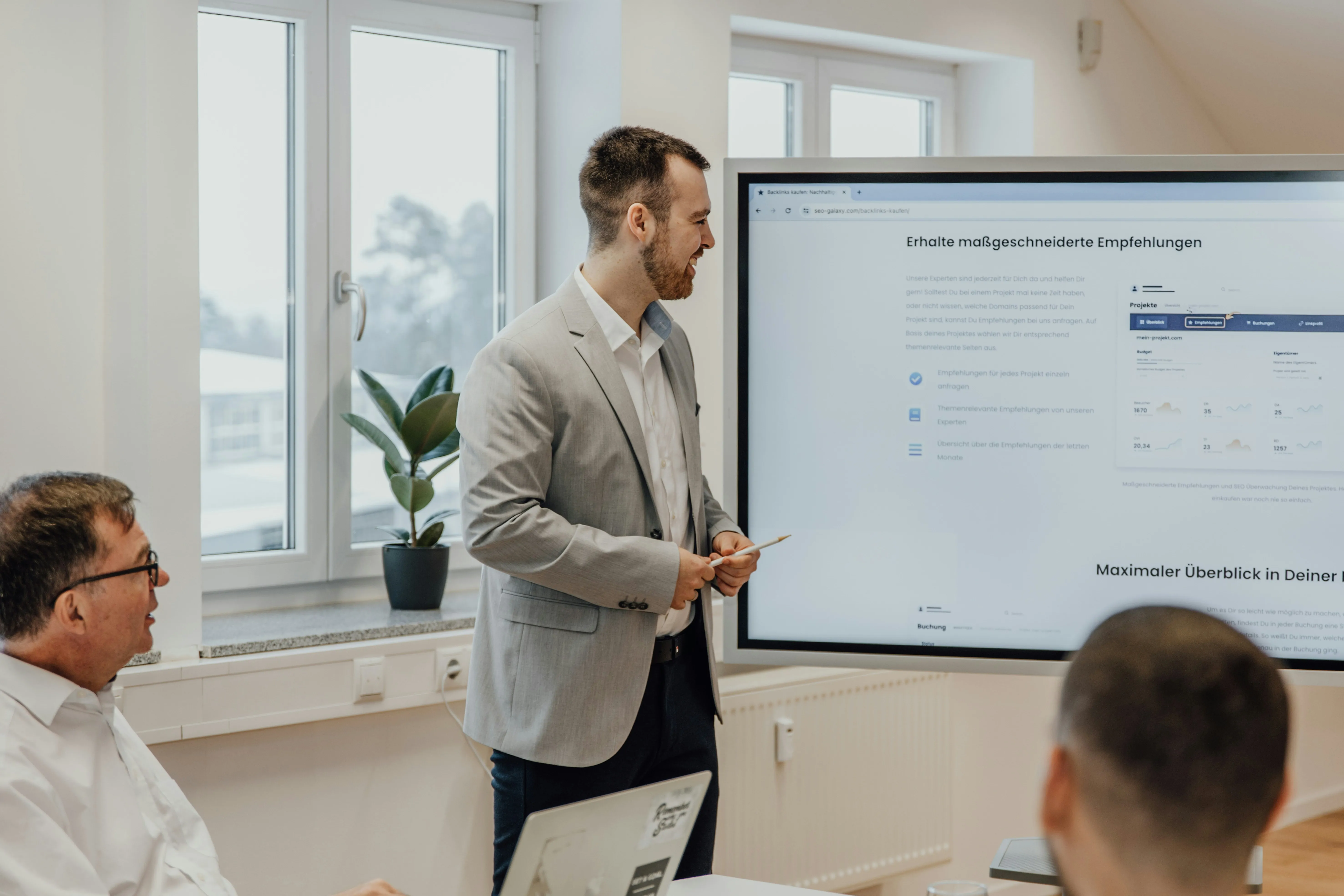 A man in a gray suit presenting in front of a large screen showing a website with German text to two seated colleagues in a bright office room.