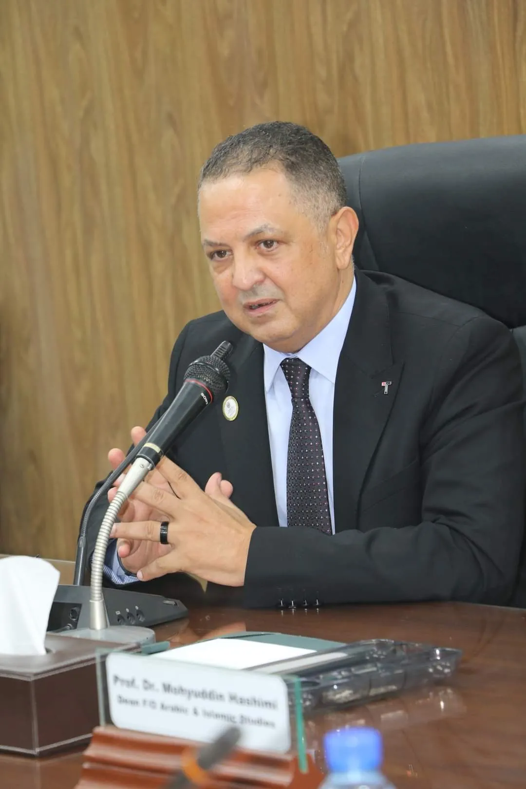 Man in a black suit and dotted tie speaking into a microphone at a desk with a nameplate and tissue box.