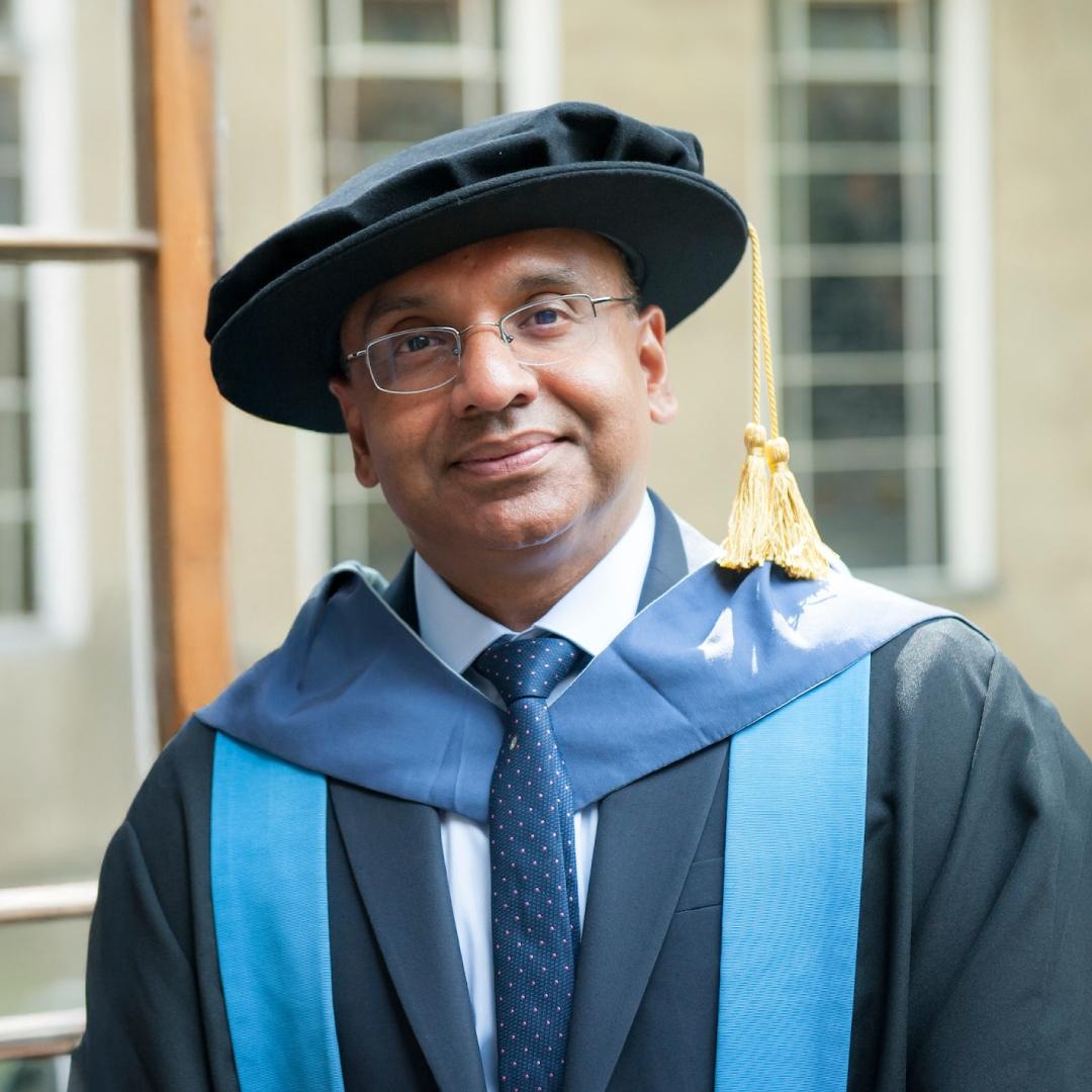 Man in a black suit and dotted tie speaking into a microphone at a desk with a nameplate and tissue box.