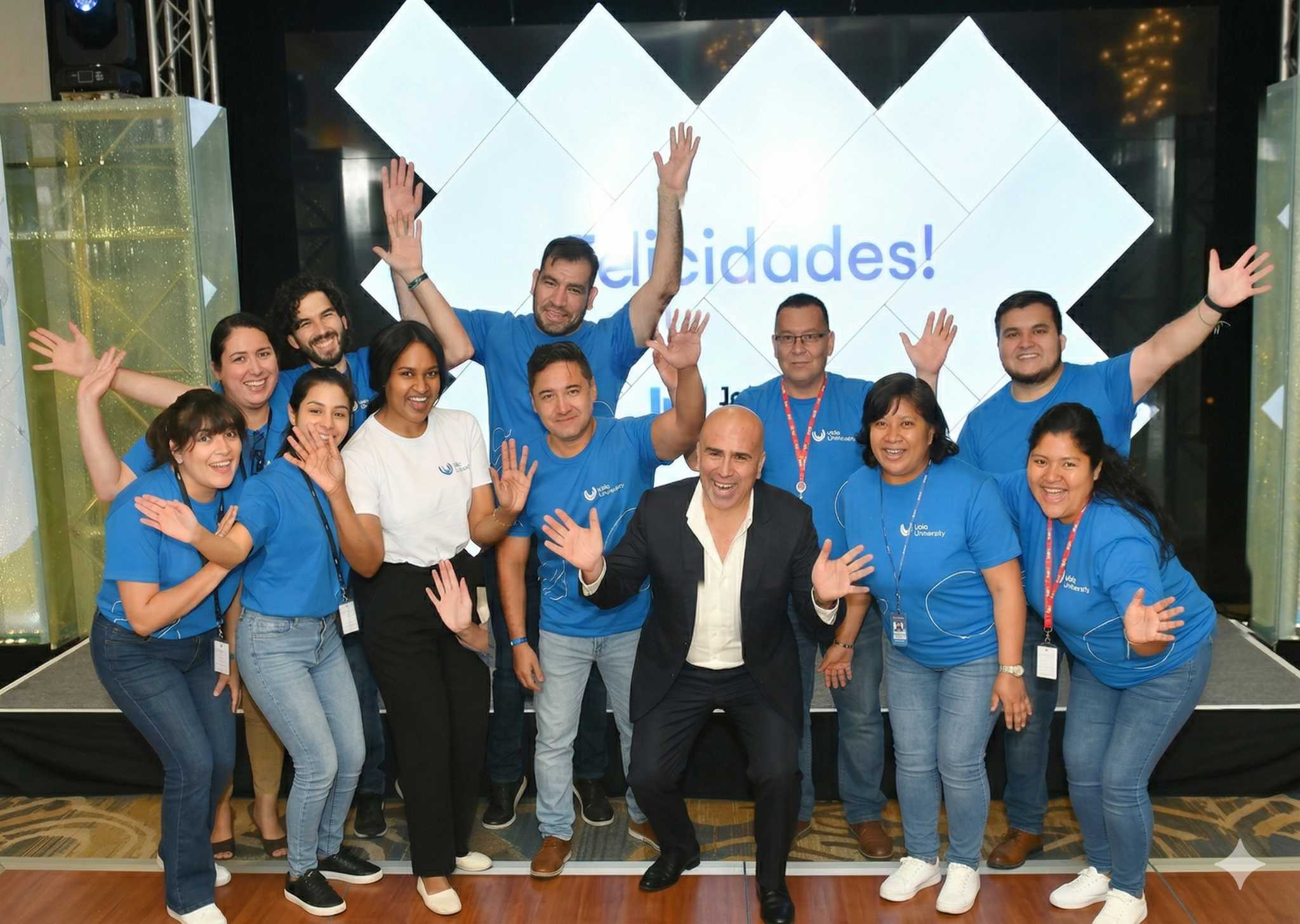 Group of smiling people waving, most in blue Jala University shirts, in front of a stage screen.