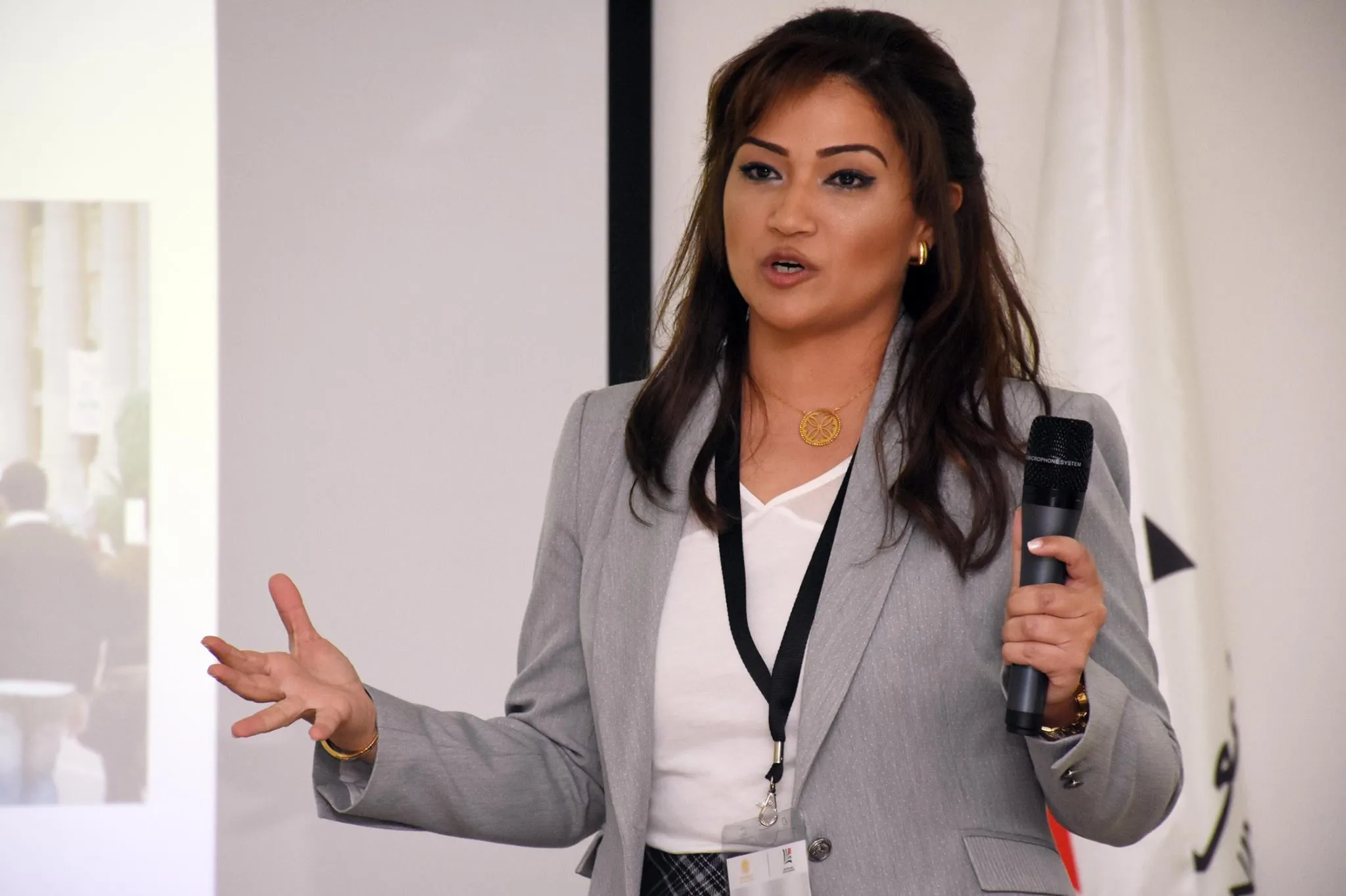 Woman in gray blazer holding a microphone speaking and gesturing during a presentation.