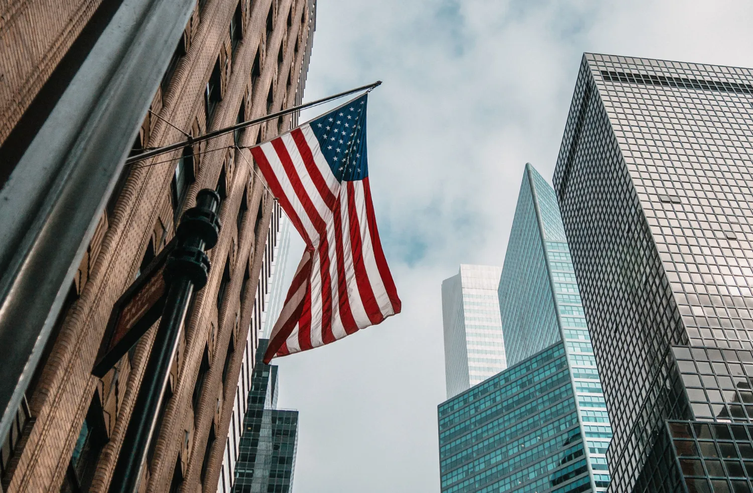 United States flag hanging from brick building with modern skyscrapers and cloudy sky background.