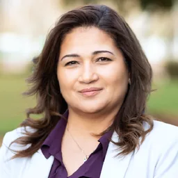 Headshot of a woman with shoulder-length hair wearing a white blazer and purple top outdoors.