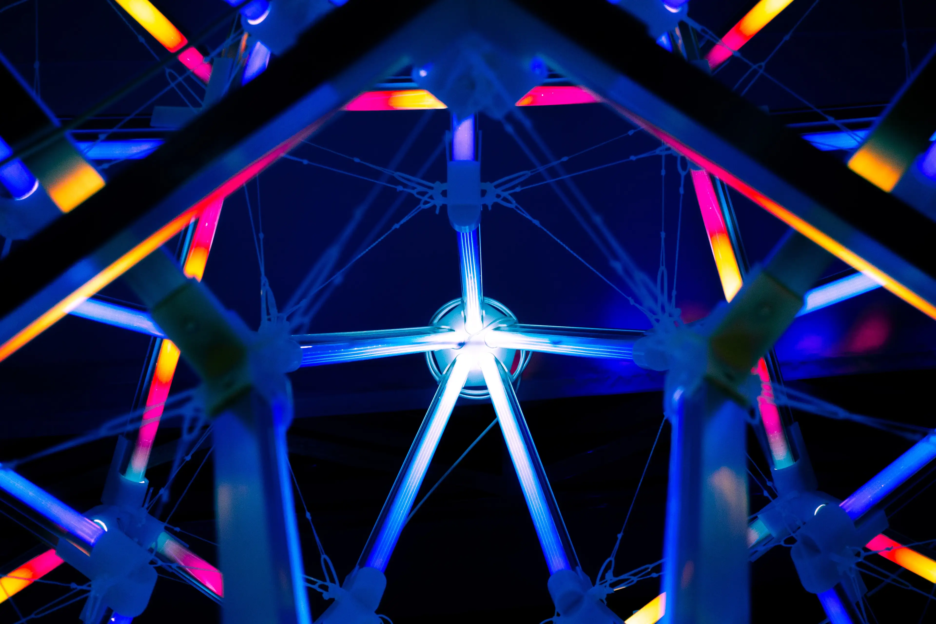 Close-up view of glowing neon tubes in blue, pink, and orange arranged in a geometric structure against a dark background.