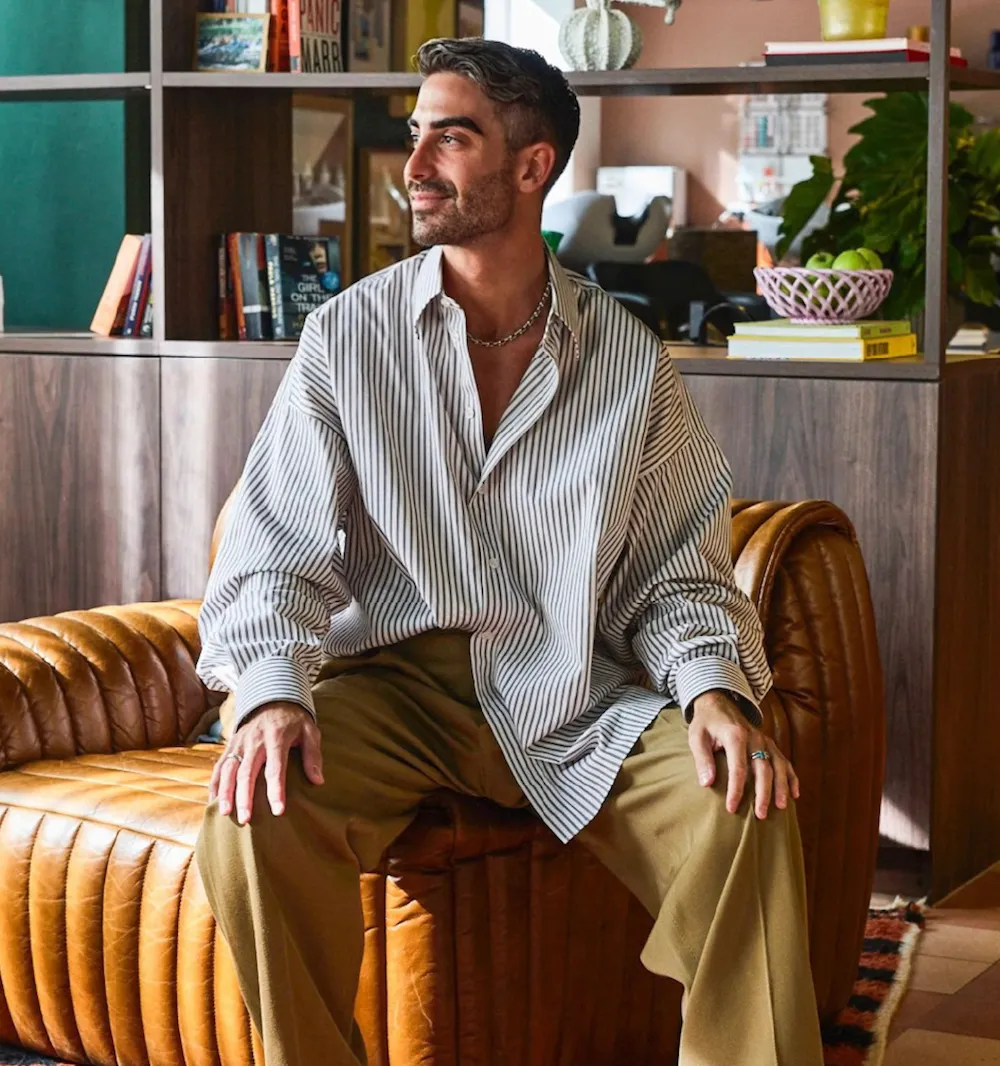 Man with dark hair and gray highlights wearing a striped shirt and brown pants sitting on a tan leather sofa in a cozy room with bookshelves and plants.