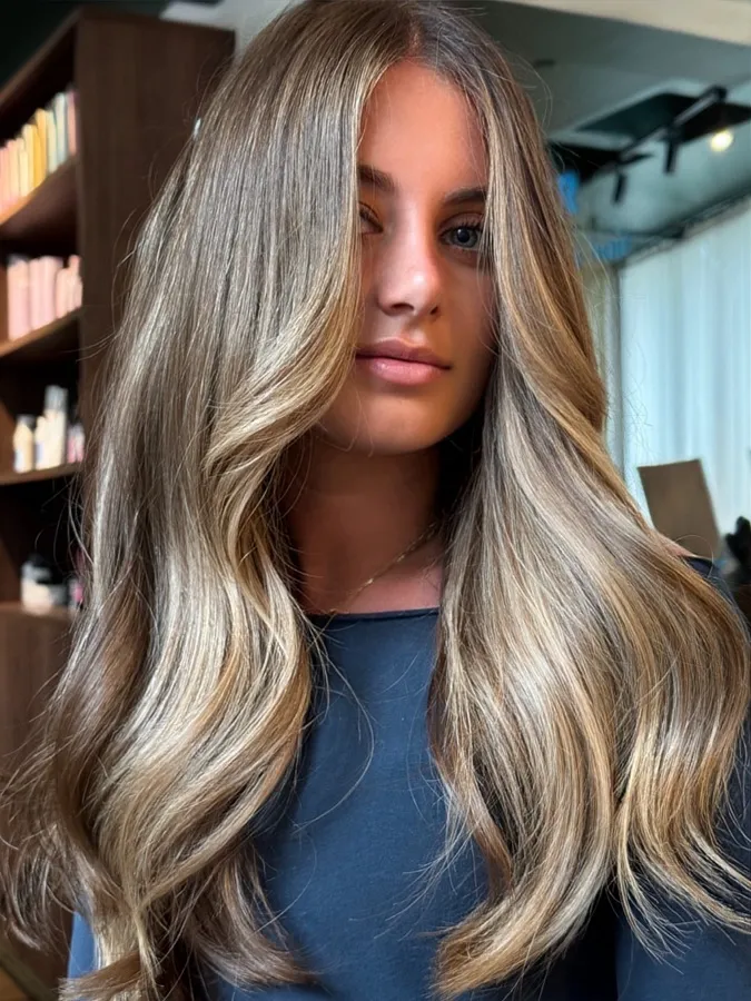 Woman with long, voluminous, wavy blonde hair and subtle highlights, wearing a dark top indoors.