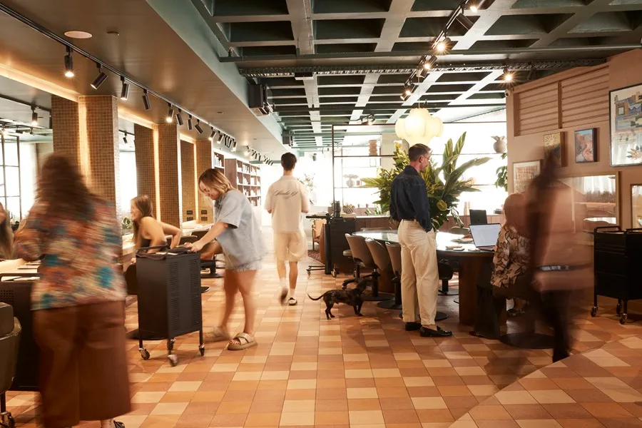People working and moving inside a modern office space with patterned tile flooring, plants, and computers, including a small dog walking near a man in beige pants.