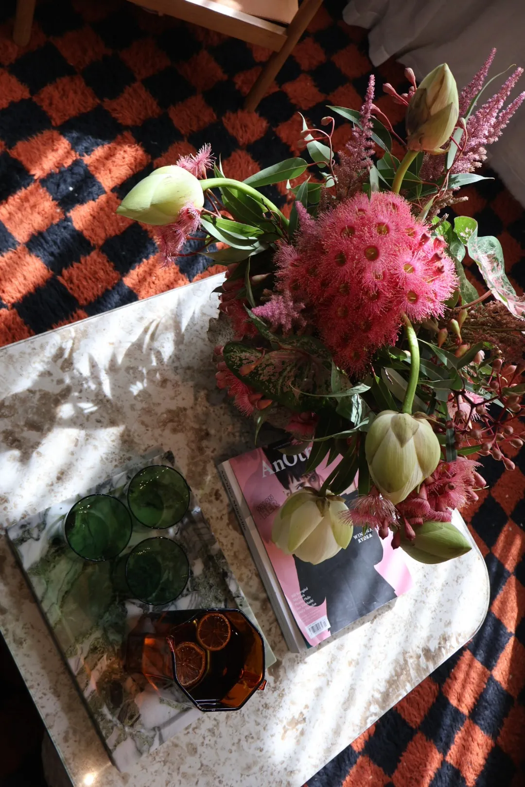 Table with a bouquet of pink and green flowers, green glasses on a marble tray, and a pink magazine on a patterned orange and black rug.