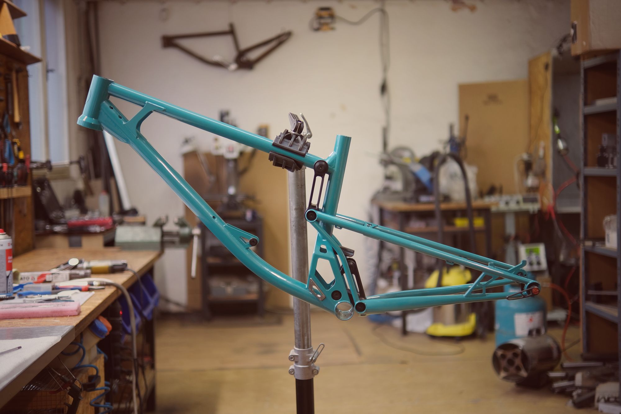Teal mountain bike frame mounted on a repair stand in a workshop with tools and equipment around.