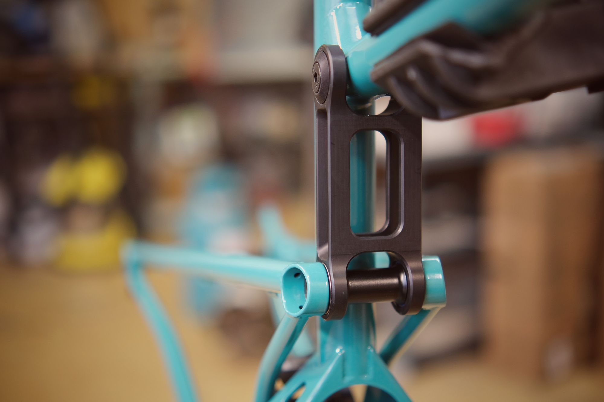 Close-up of a hand wearing a glove assembling the rear suspension linkage on a turquoise bicycle frame in a workshop.