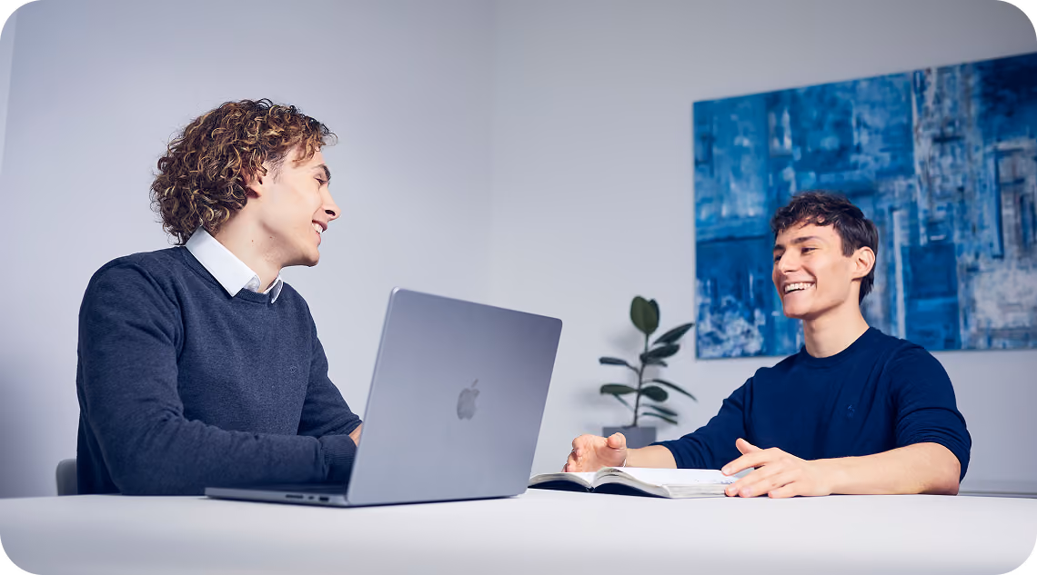 Emil und Elia, die Gründer von THEFABERS, lachen bei einer Besprechung am Tisch mit MacBook und Notizbuch
