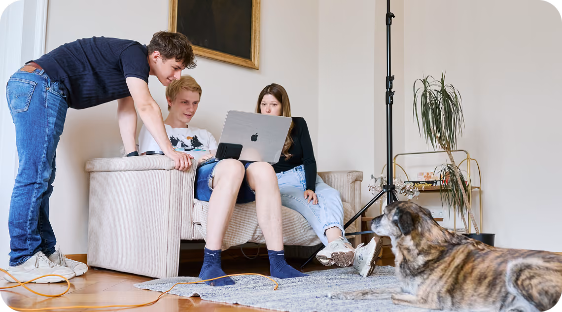 Elia, Matthias und Anna schauen beim Fotoshooting gemeinsam auf ein MacBook, im Vordergrund liegt ein Hund